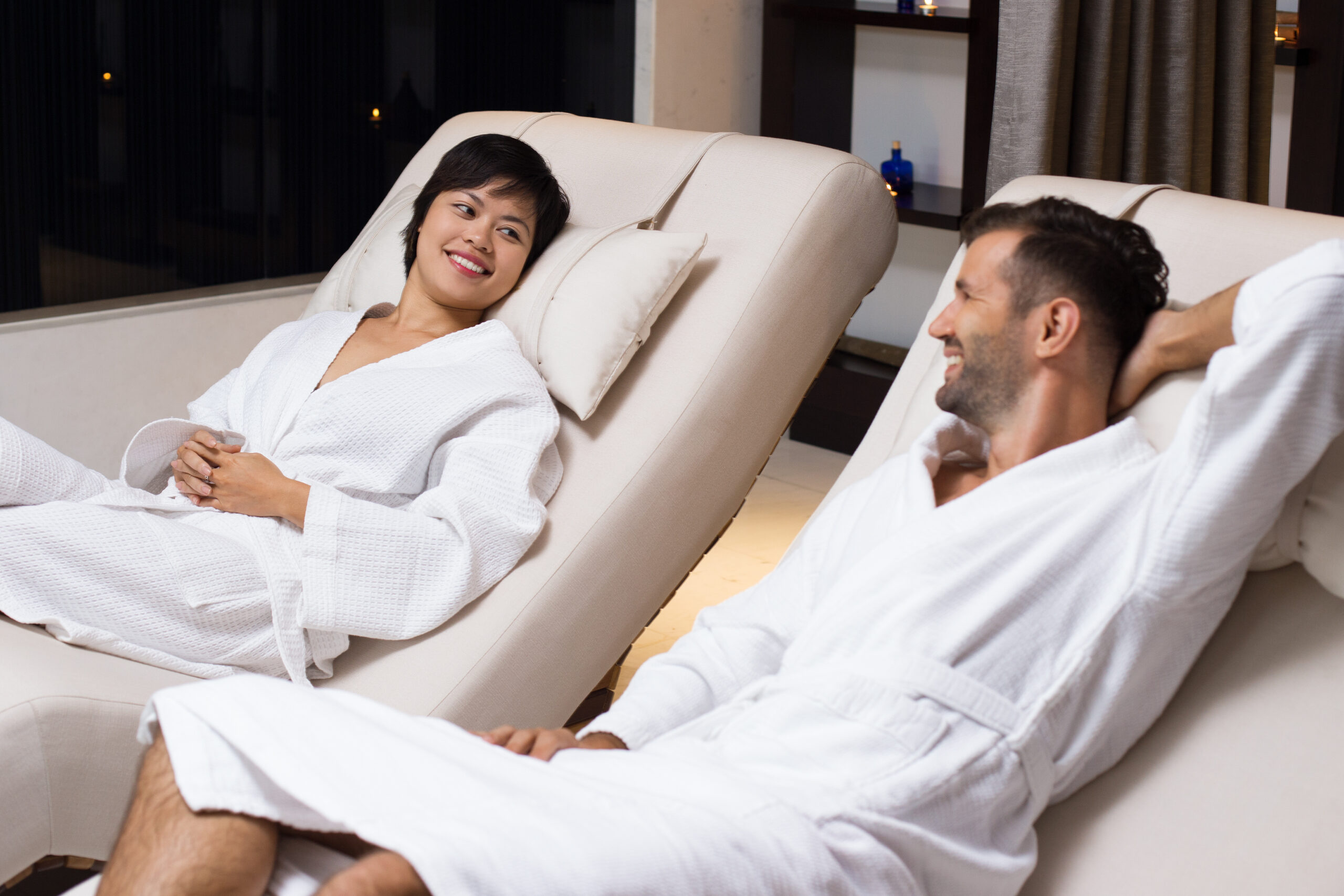 Smiling young woman and man wearing bathrobes, chatting and lying on deckchairs in spa salon